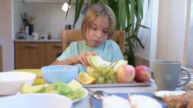 Young boy sitting at the kitchen table eating fresh fruits including orange, grapes, melon, and apples.