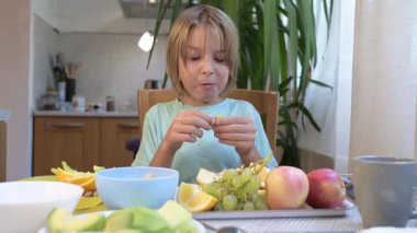 Young boy sitting at the kitchen table eating fresh fruits including orange, grapes, melon, and apples.