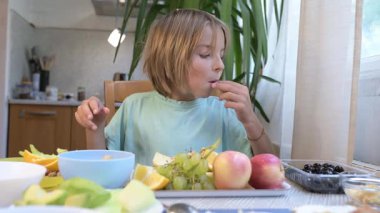 Young boy sitting at the kitchen table eating black olives with a tray of fresh fruits including apples, grapes, and oranges.