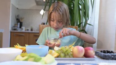 Young boy sitting at the kitchen table eating fried egg from a glass bowl with fruits like apples, grapes, and oranges on the table.