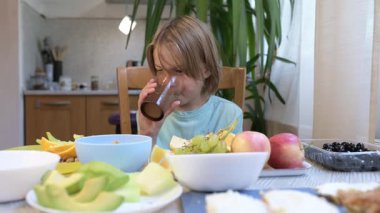 Young boy drinking lemonade at a kitchen table, surrounded by fresh fruits and avocado slices, enjoying a healthy snack at home.