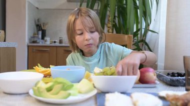 Young boy sitting at the kitchen table holding a blueberry with a bowl of berries and a plate of sushi rolls in front of him.