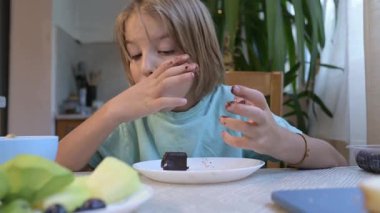 Young boy eating chocolate dessert bar at the kitchen table, surrounded by fresh fruits and avocado slices, enjoying a sweet snack at home.
