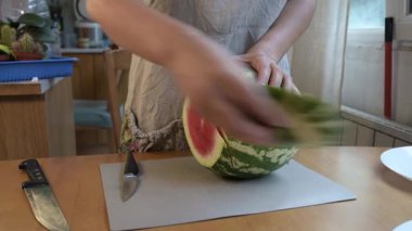 Hands slicing a large striped watermelon on a cutting board in a home kitchen, preparing fresh fruit for eating.