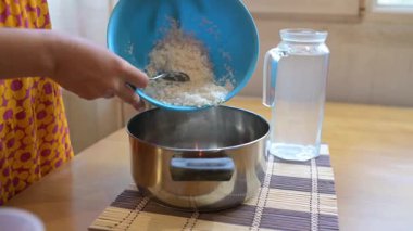 Close-up video of a person pouring washed rice from a blue bowl into a metal pot on a wooden table, preparing to cook rice at home.