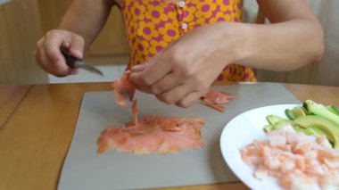 Close-up video of a woman slicing smoked salmon on a cutting board with avocado and shrimp pieces on a plate nearby, preparing sushi ingredients.