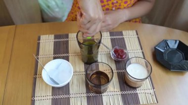 Close-up video of a woman preparing matcha latte at home, mixing ingredients in glasses on a wooden table with ice, milk, and matcha tea.