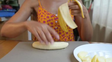 Close-up video of a woman slicing a banana with a knife on a cutting board in the kitchen, preparing fresh fruit for a healthy meal or dessert.