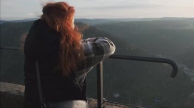 Kızıl saçlı kız düşünceli bir şekilde dağlara bakıyor. Rüzgarda uçuşan saçlar gün batımına karşı. Yüksek kalite 4k görüntü