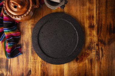 Tahta kırsal masa üzerinde boş taş comal plaka, kil çömlek ve taş molcajete ile renkli tipik Meksika kumaşı, Meksika gastronomisinde geleneksel mutfak gereçleri.