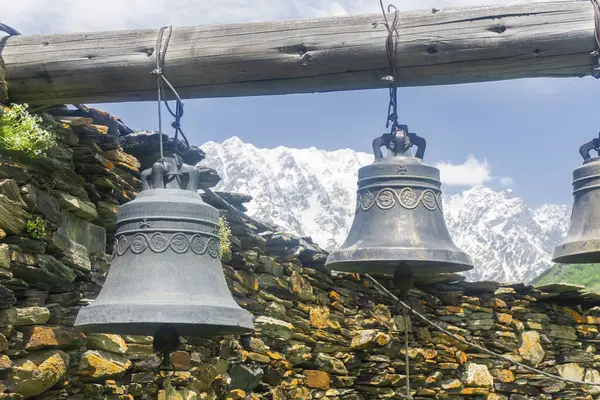 stock image Two bells hang on a wooden log near a stone wall in the courtyard of the Lamaria Monastery in the village of Ushguli. The snow capped peaks of Mount Shkhara are in the background.