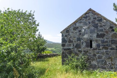 Ortaçağ kilisesinin avlusunda. Taş duvar, küçük pencere. Ağaçlar, çalılar ve parlak mavi gökyüzü