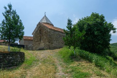 Kiliseye giden toprak yol. Taş duvar ve kubbe görünür durumda. Ağaçlar ve çalılar var. Bulutlu parlak mavi gökyüzü. Vejini köyünün Amagleba Kilisesi, Georgi