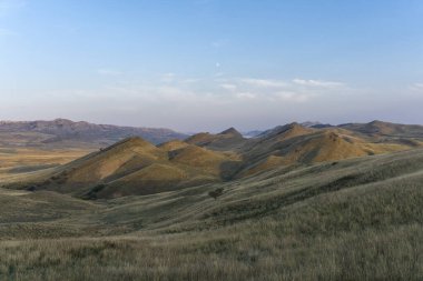 Dağ savanası manzarası. Dağ tepeleri görülebilir. Kuru yeşil ve turuncu çimenler. Bulutlu açık mavi gökyüzü