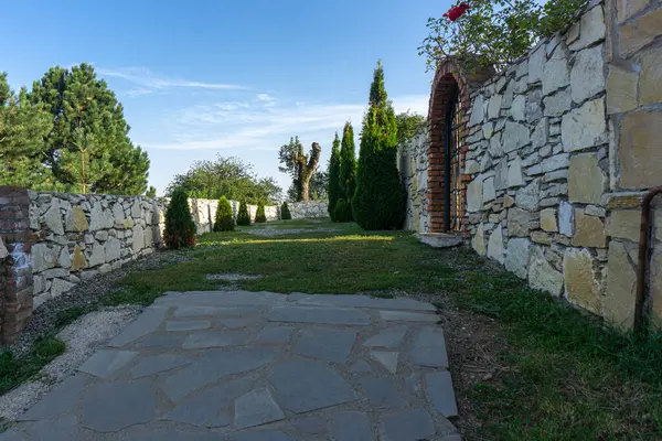 Taş ve çimen yolu avluya çıkıyor. Taş bir duvarda tuğla kemerli desenleri olan güzel demir kapı. Kozalaklı ağaçlar. Georgia, Udzo Manastırı.