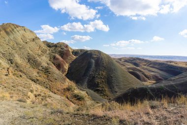 Georgia 'daki Mravaltskaro Barajı yakınlarında çok renkli dağlar. Kayaların yüzeyinde gri, yeşil, kırmızı ve turuncu toprak şeritleri. Bulutlu parlak mavi gökyüzü. Kuru ot.