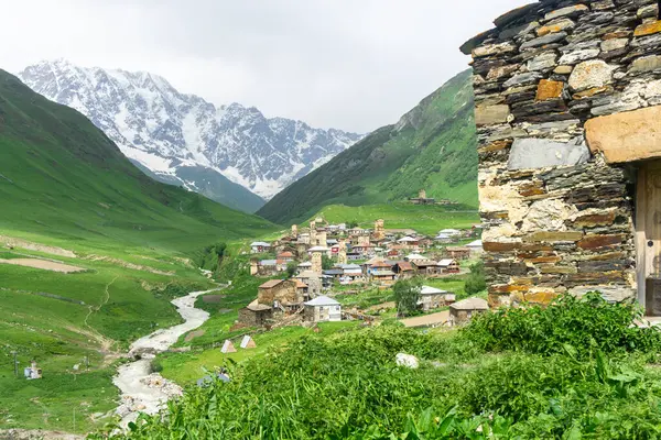 stock image The wall of a stone church on a hill, the village of Ushguli in a gorge between the mountains. The flowing river and the snow capped peaks of Mount Shkhara