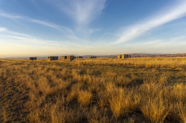 Evening view of an earthen road through the savannah and an abandoned village. Bright blue sky with clouds, orange grass and unfinished houses. Low mountains around. Georgia. clipart