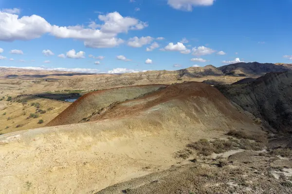 Georgia 'daki Mravaltskaro Barajı yakınlarında çok renkli dağlar. Kayaların yüzeyinde gri, yeşil, kırmızı ve turuncu toprak şeritleri. Bulutlu parlak mavi gökyüzü. Kuru ot.