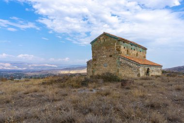 Tepedeki Pavnisi Kilisesi ve manzara manzarası vadi ve dağlar. Turuncu döşeme ve duvarda renkli tuğlalar. Parlak mavi gökyüzü ve bulutlar. Mezar taşları görünür. Georgia.
