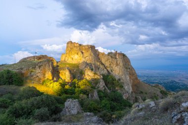 Sıcak ışıkla Kojori kalesinin akşam aydınlatması. Şiddetli bulutlar, dağlar ve arka planda bir köy olan vadi