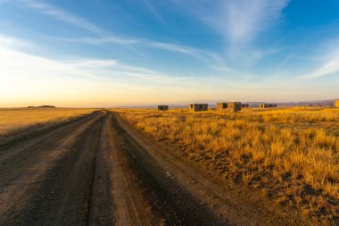 Evening view of an earthen road through the savannah and an abandoned village. Bright blue sky with clouds, orange grass and unfinished houses. Low mountains around. Georgia. clipart