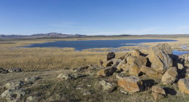 Kocaman taşlar ve berrak suyla dolu bir göl. Dağlar ve tarım alanları. Parlak mavi gökyüzü