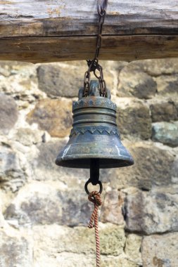 Süslü eski bir bronz çan zincirli bir kütüğe asılı. Arkaplanda bulanık taş duvar