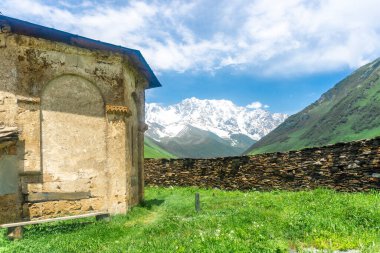 Lamaria Manastırı 'nın taş kilisesinin köşesinde. Bahçenin yüksek taş duvarı. Shkhara Dağı 'nın karlı zirveleri. Bulutlu parlak gökyüzü