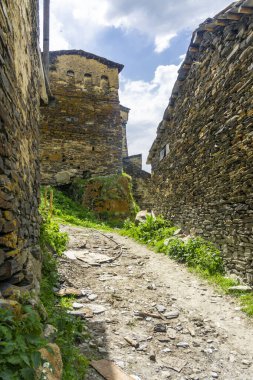 Yüksek taş duvarlar arasında dik bir toprak yol. Ushguli köyünün kale kuleleri ve caddeleri.