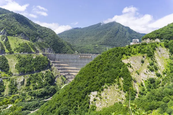 stock image Enguri reservoir dam. Tree covered hill with approach roads. Mountains and bright sky in the background