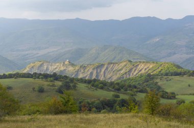 Uçurumun üstündeki küçük kilise güneş ışığıyla aydınlanır. Önde yeşil ağaçlar var. Arka planda dağlar ve gökyüzü