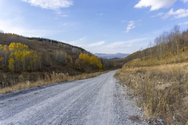 The gravel road runs between the hillocks. Beautiful autumn trees and bushes. Bright blue sky with clouds clipart