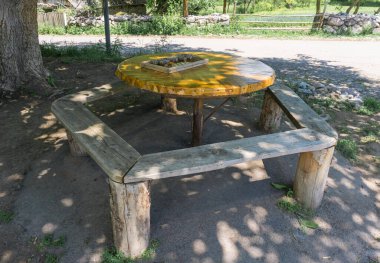 Yuvarlak masa ve bir ağacın gölgesinde ahşap banklar. Dominolar ve abaküs masada.