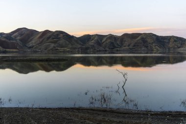Rezervuarda güneş doğmadan önce. Mavi gökyüzü ve dağlar suya yansıyor. Kırmızı bulut. Kuru dallar sudan çıkıyor.