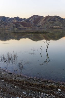 Rezervuarda güneş doğmadan önce. Mavi gökyüzü ve dağlar suya yansıyor. Kuru dallar sudan çıkıyor.