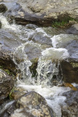 Kayaların üzerinde çağlayan bir şelalenin yakın görüntüsü. Su damlaları, kuru yapraklar ve köpük görülebilir.