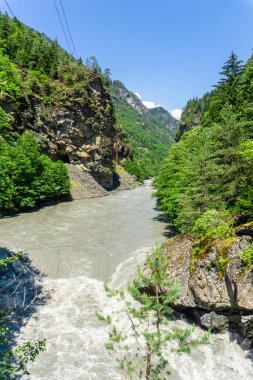 Çamurlu bir dağ nehri ağaçlar, kayalar ve çalılar arasında akar. Nehir yatağında büyük taşlar var. Yüksek gerilim kabloları görünür.