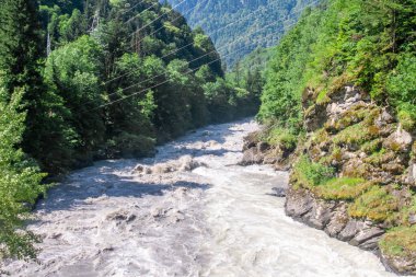 Çamurlu bir dağ nehri ağaçlar, kayalar ve çalılar arasında akar. Nehir yatağında büyük taşlar var. Yüksek voltaj direkleri ve kablolar görülebilir.