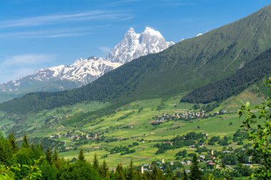 Orman ve çalılarla kaplı dağlar. Vadideki köyler. Ushba Dağı 'nın karlı zirvesinin üzerinde hafif bulutlar