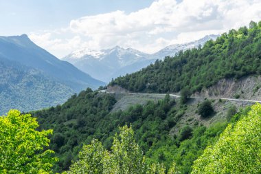 Dağ yolu uçurum ve vadi arasından geçer. Orman ve çalılar. Arka planda karlı dağ zirveleri ve uçurumlar var.