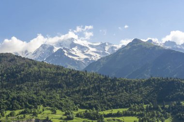Karla kaplı dağ zirveleri bulut seviyesinde. Önplanda ağaçlarla kaplı tepeler