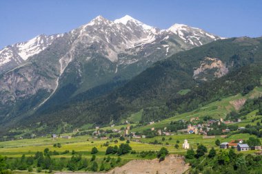 Dağın altındaki vadide bir köy. Kilise ve savunma kuleleri görünür durumda. Arka planda karla kaplı dağlar