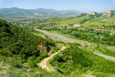 Nehir boyunca uzanan Ksani Nehri 'nin üzerinden, çim ve çalılarla kaplı düzlüklere bakın. Arka planda dağlar ve mavi gökyüzü