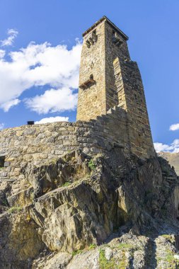 Uçurumun kenarında duran Sno köyü kalesi. Taş duvar ve yüksek kule. Dağlar, arkasında bulutlar olan mavi gökyüzü.