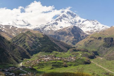 Dağların altındaki Gergeti köyü, tepedeki Gergeti Trinity Kilisesi ve kar kaplı Mkinvartsveri Kazbek. Bulutlu mavi gökyüzü
