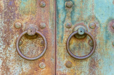Paslı metal kapılardaki iki tokmağı yakından izle. Oval saplar, perçinler. Kırmızı ve turkuaz renkler