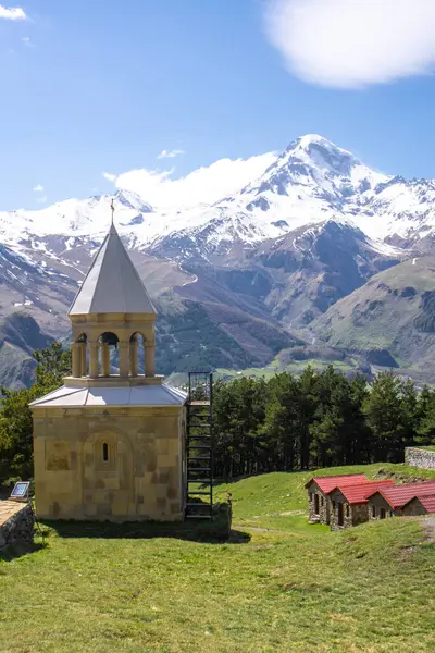 stock image The Ioane Natlismcemeli monastery bell tower, monks' houses and snow capped Mount Mkinvartsveri Kazbek. Coniferous trees, blue sky with clouds