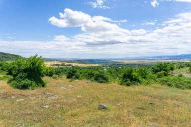 Sarı çimenli, yeşil çalılı, dağlı, vadili, kanyonlu. Parlak mavi gökyüzü.