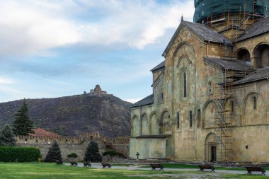 Svetitskhoveli Katedrali avlusu. Kilisenin sol duvarı, yeşil çimenler, banklar, koltuklar. Arka plandaki tepedeki Jvari Manastırı. Parlak gökyüzü